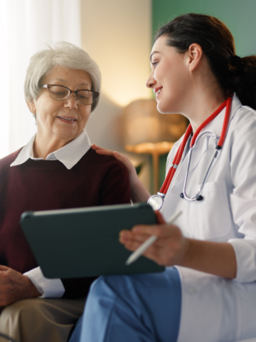 A care provider in a lab coat, with a stethoscope, sits on a sofa with an older woman wearing a burgundy sweater with a white collared shirt. The two of them are looking at each other and having a conversation while the care provider shows the lady a tablet screen.