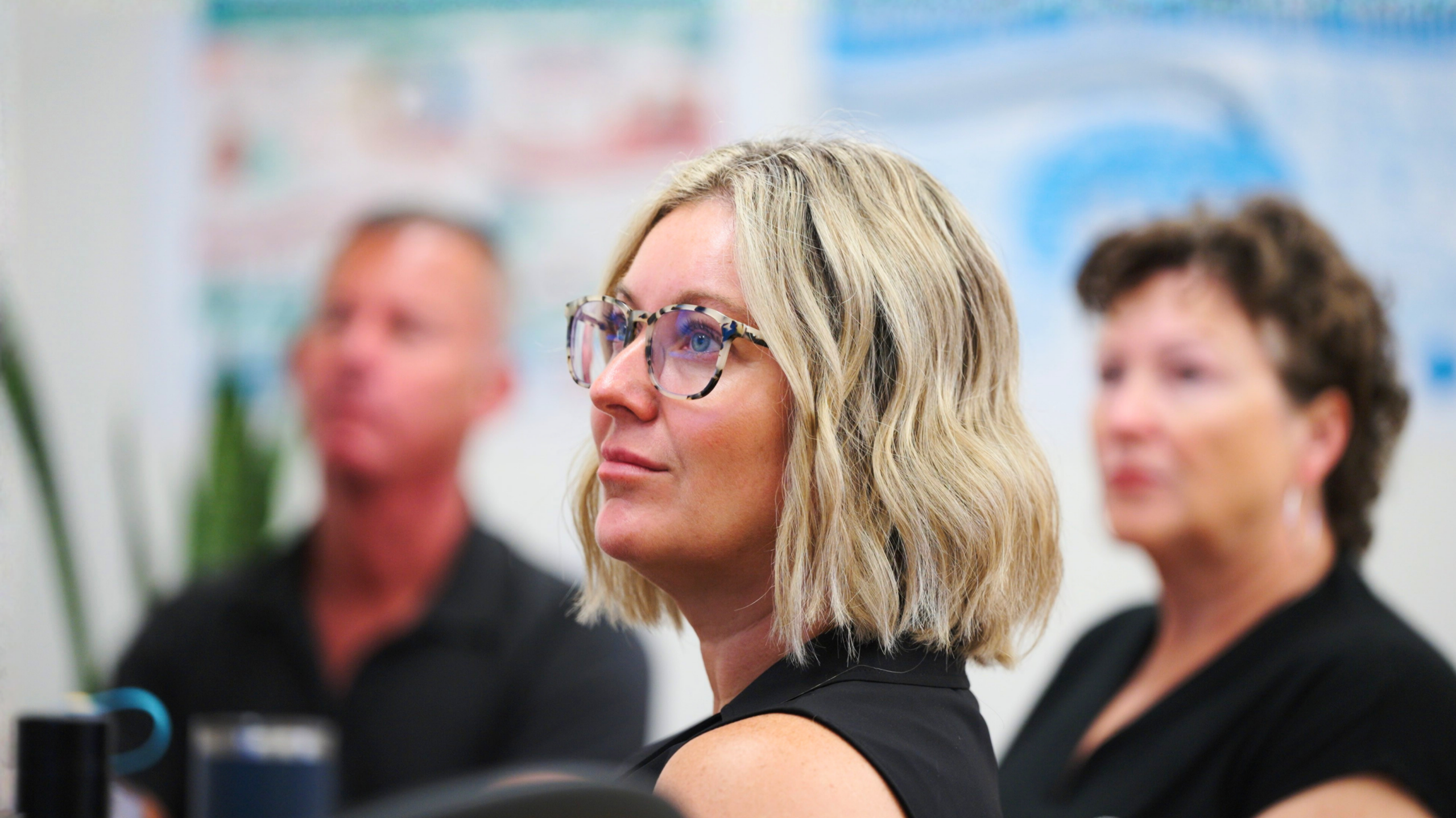 A blonde woman wearing animal print glasses in the foreground with an out of focus man and woman behind her.